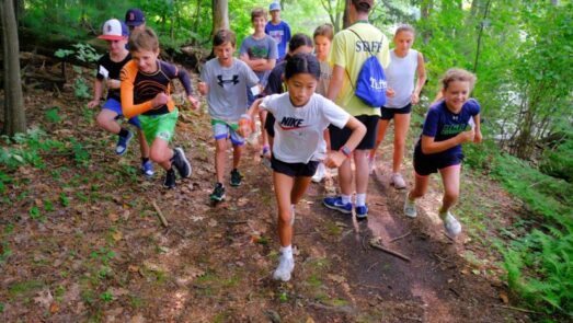 A group of children and an adult wearing a "staff" shirt are running up a forested trail.